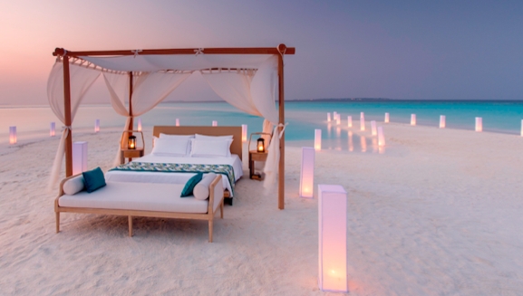 A canopied four-poster bed draped in muslin surrounded by candles on a sandbank in the Maldives