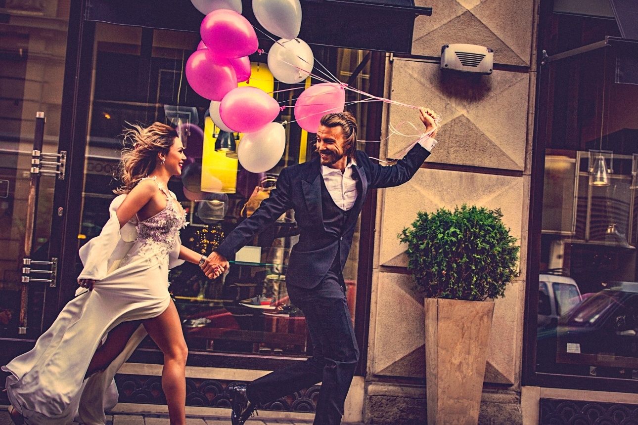 man in suit woman in smart dress running in street holding a bunch of balloons