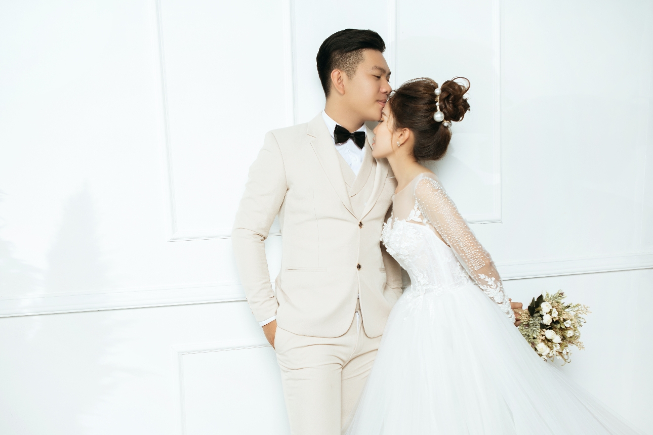 bride and groom standing up against a white wall with the groom kissing the brides forehead