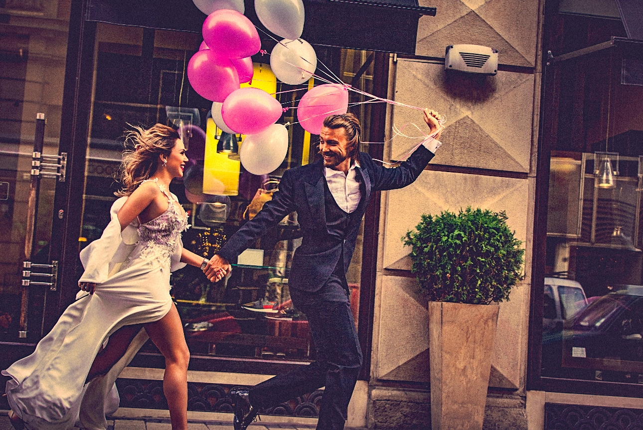 bride and groom in modern clothes running through city street with a bunch of pink ballons