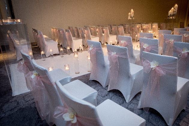 White chairs in rows with pink bows on sitting next to a while aisle runner decorated with candles