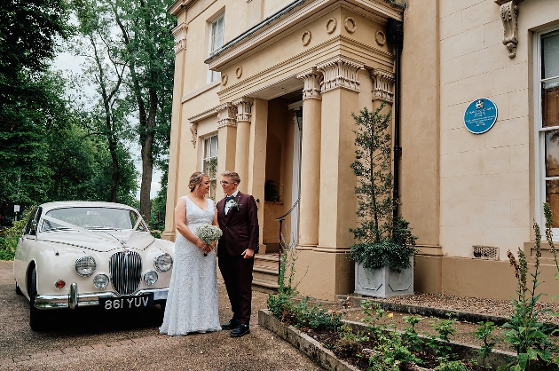 A bride and groom standing next to a cream car outside a grand manor house
