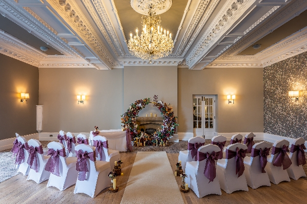 A round floral arch at the end of an aisle that's flanked by white chairs with purple bows on
