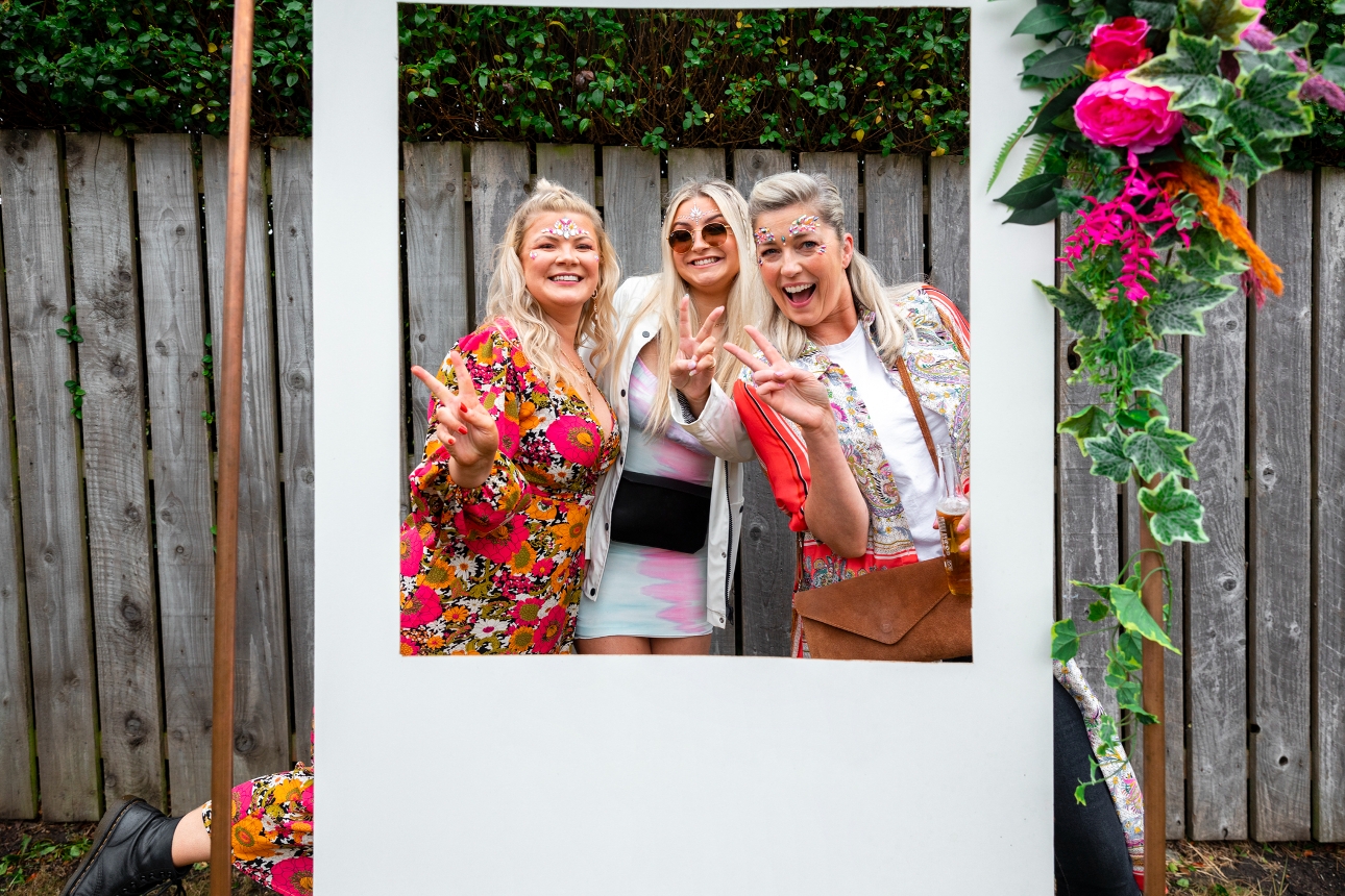 three friends dresses up for festival stood in front of picture frame for photo
