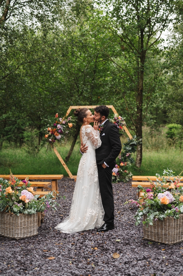 A bride and groom embracing in front of a wooden ceremony arch