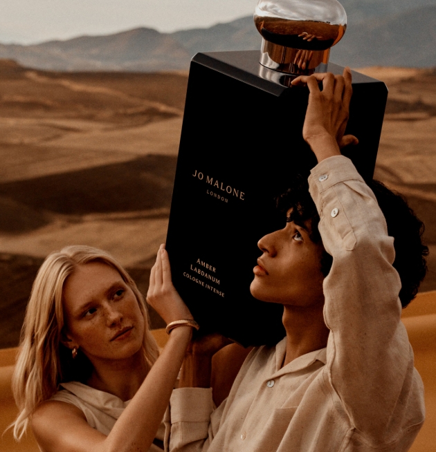 man and woman in the dessert holding a massive perfume bottle